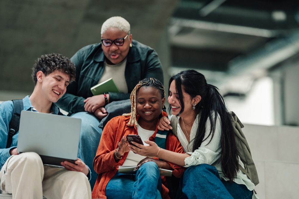 Multi-ethnic college students laughing and studying together indoors, sharing laptops and phones, connecting and collaborating on campus